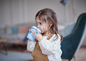 Ein Mädchen trinkt Milch aus einer Schnabeltasse. Im Hintergrund sieht man ein Wohnzimmer.