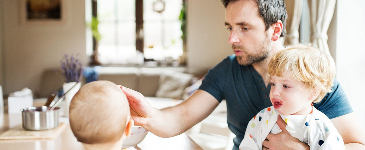 Vater mit Kleinkind auf dem Schoss füttert ein Baby.