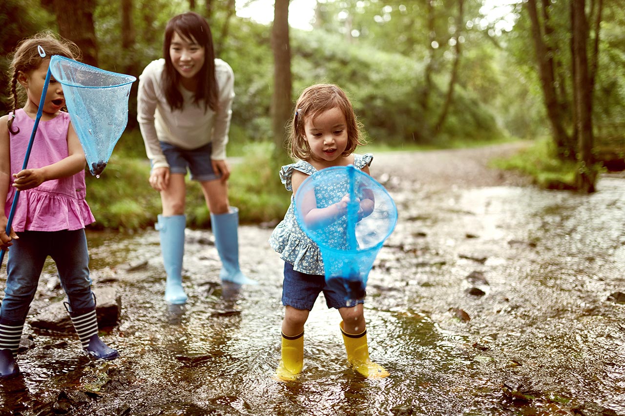 Mama mit zwei Kleindkindern spielt draußen im Matsch. Beide Mädchen haben jeweils einen Kescher in der Hand. Im Hintergrund sieht man einen Wald.