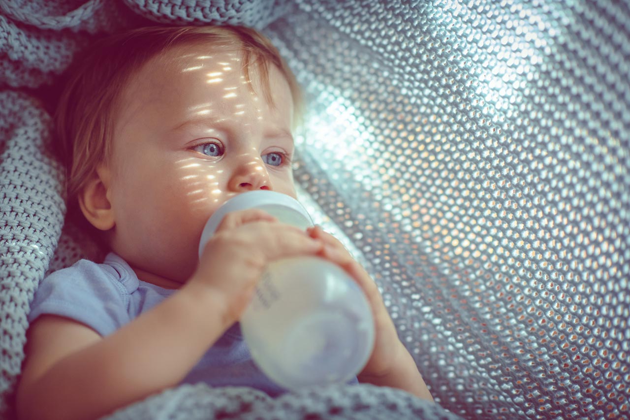 Ein Mädchen trinkt Milch aus einer Schnabeltasse. Im Hintergrund sieht man ein Wohnzimmer.