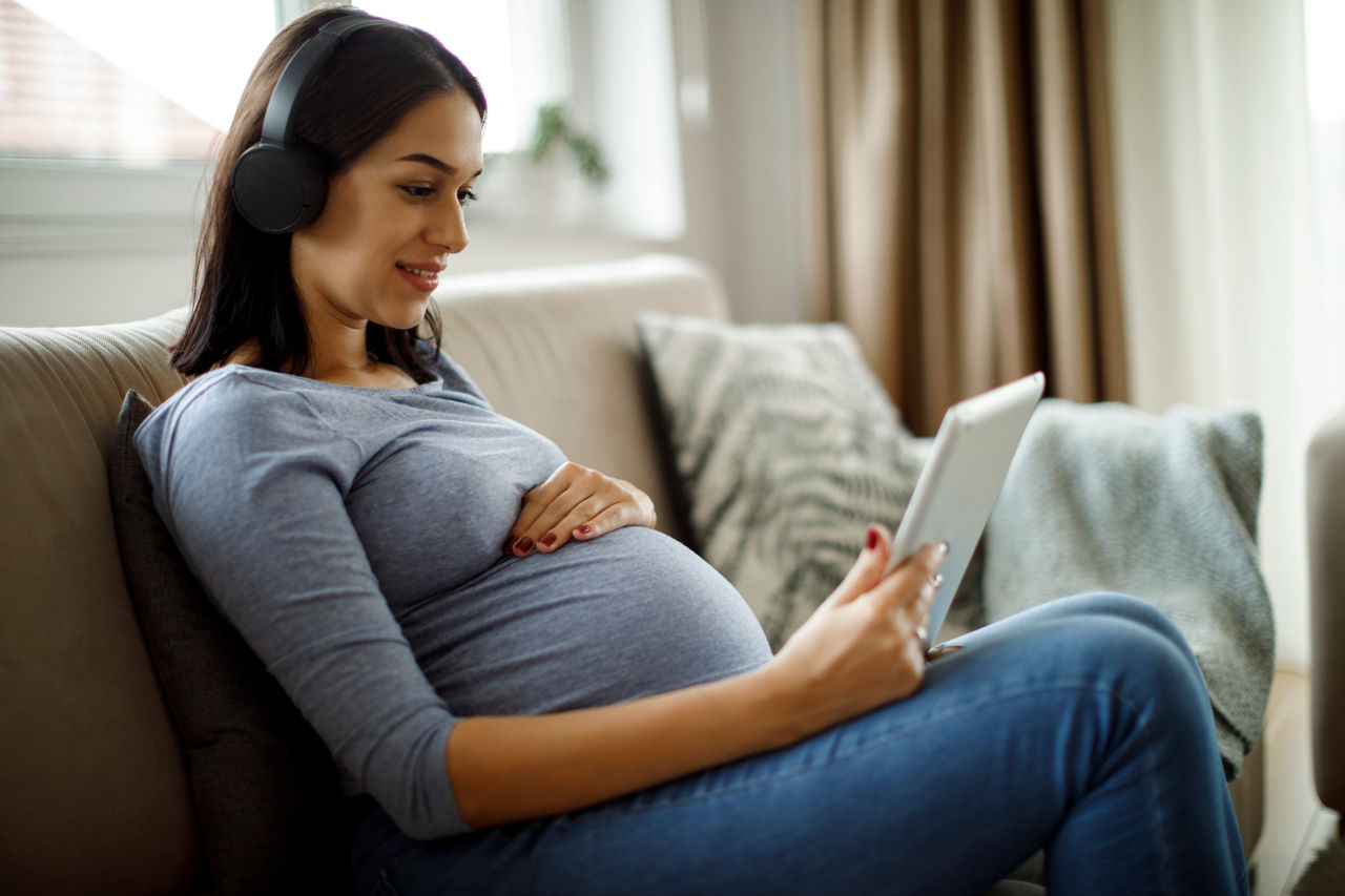 Schwangere sitzt auf der Couch mit Tablet und Kopfhörern