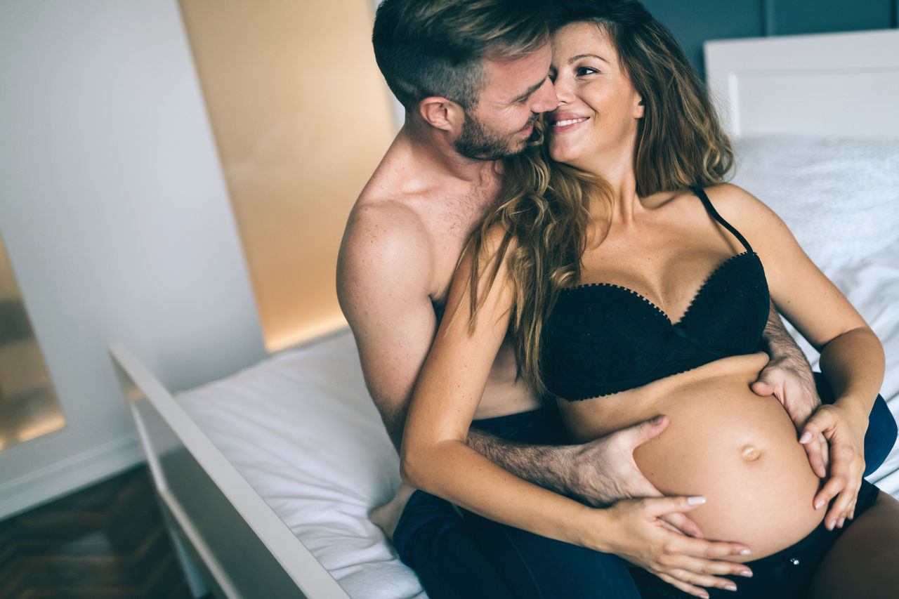 Mann und Schwangere sitzen gemeinsam auf der Couch. Der Mann hält die Frau im Arm und umfasst ihren Bauch.